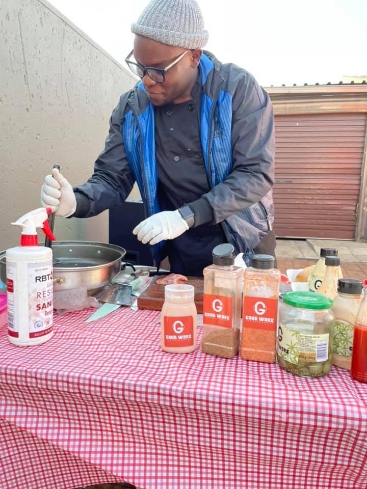 A man wearing gloves and a beanie prepares food at an outdoor table covered with spices, sauces, and sanitiser. - GrubWorx