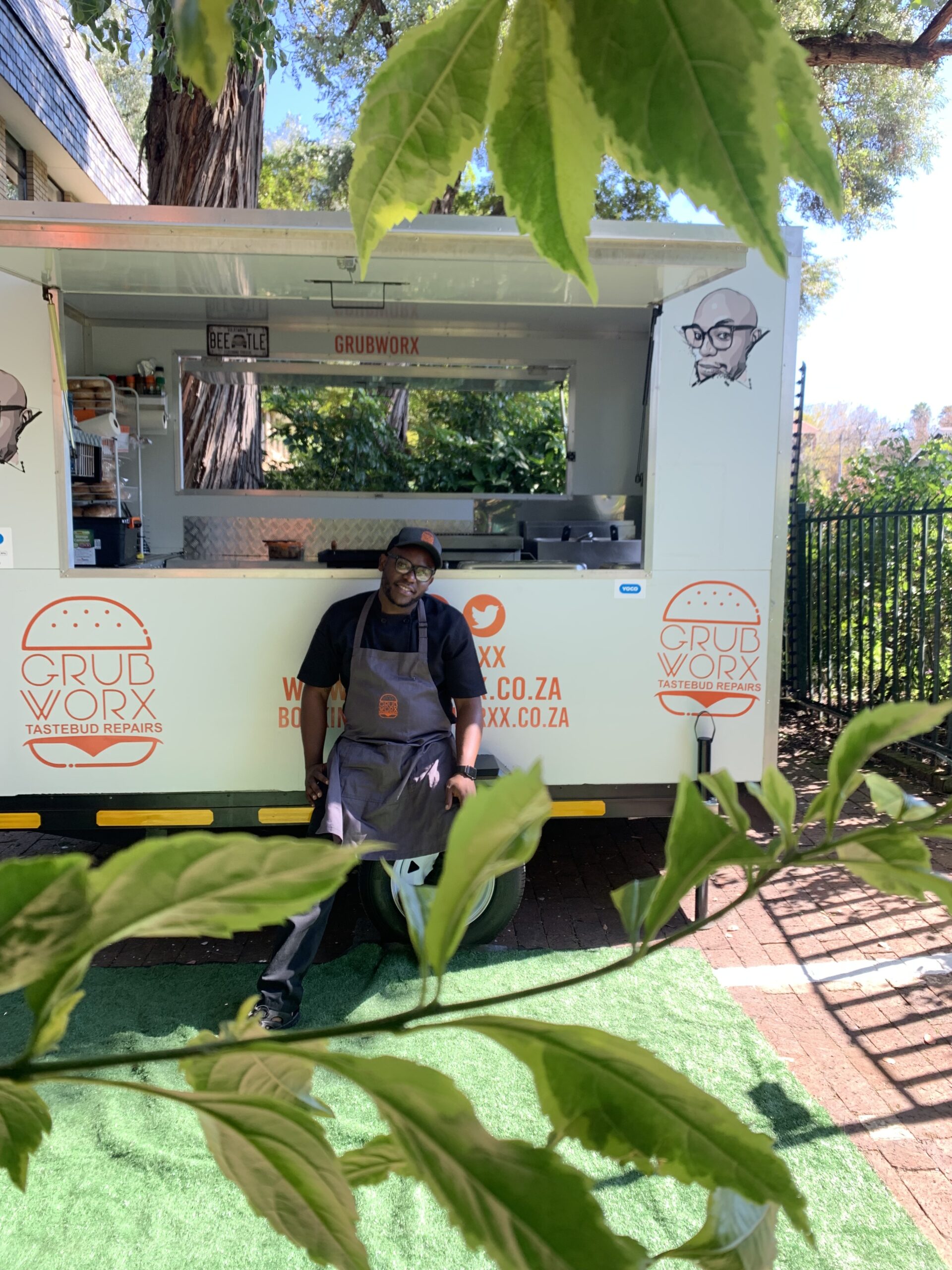A man wearing an apron stands and smiles in front of a Grubworx food van; green leaves partially frame the image in the foreground. - GrubWorx