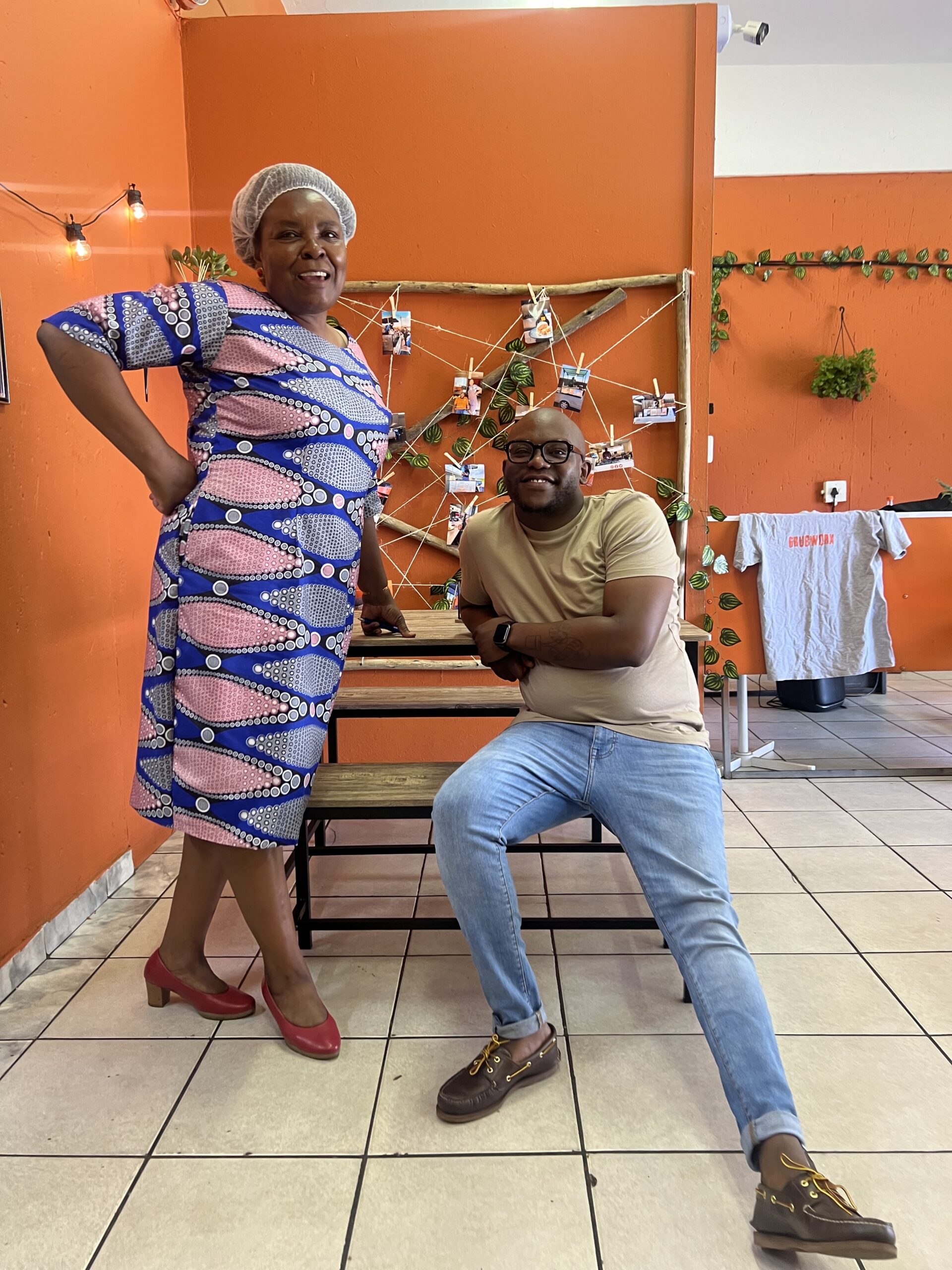 A woman in a patterned dress and hairnet stands next to a seated man in casual clothes against an orange wall decorated with photos and fairy lights. - GrubWorx