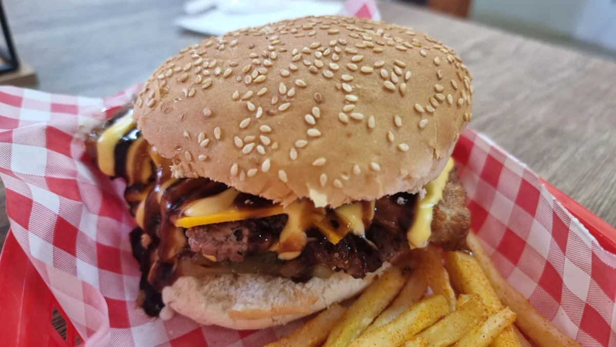 A sesame seed burger bap with a beef patty, melted cheese, and sauce, served with seasoned chips on a red and white chequered tray. - GrubWorx