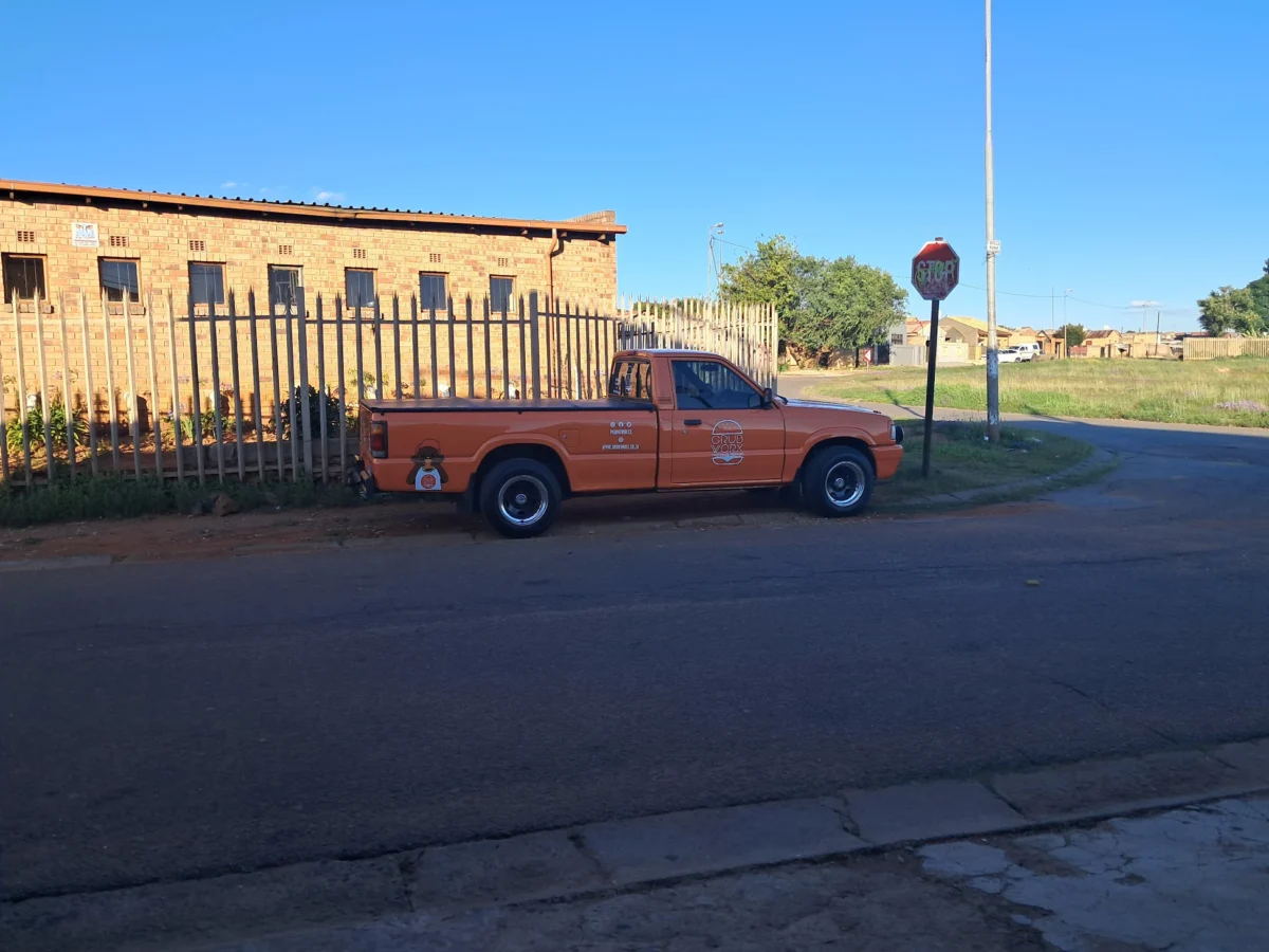 An orange pickup lorry parked on the roadside near a brick building and a stop sign on a sunny day. - GrubWorx