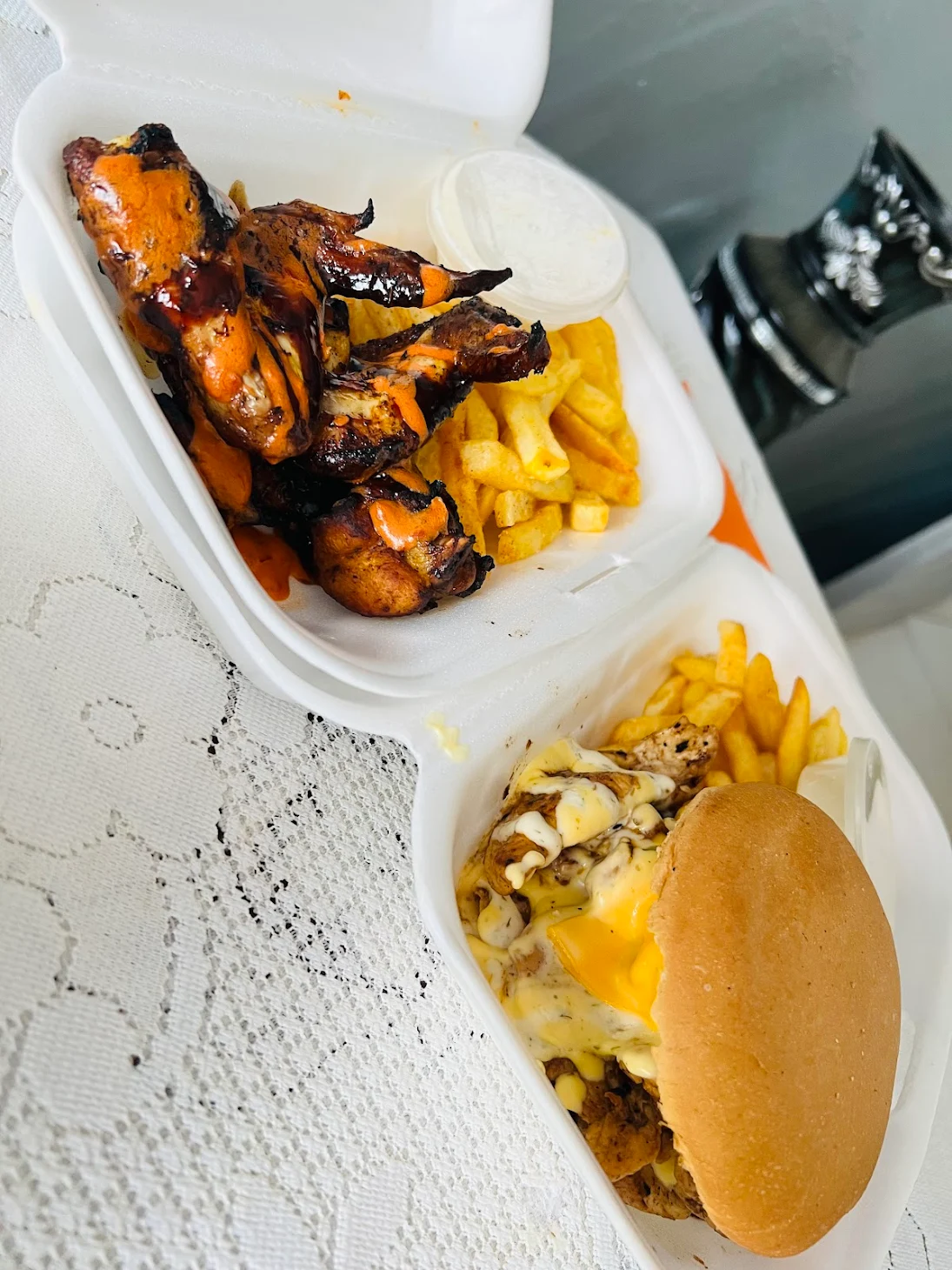 A takeaway meal in two polystyrene containers: one with sauced chicken wings, chips, and dipping sauce; the other with a cheeseburger, chips, and melted cheese. - GrubWorx